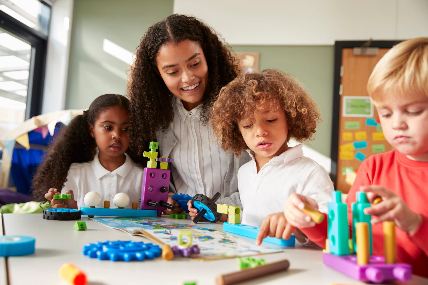 Lehrerin lächelt, während zwei Vorschulkinder (5-7 Jahre) ein technisches Spielzeug mit Anleitung zusammenbauen